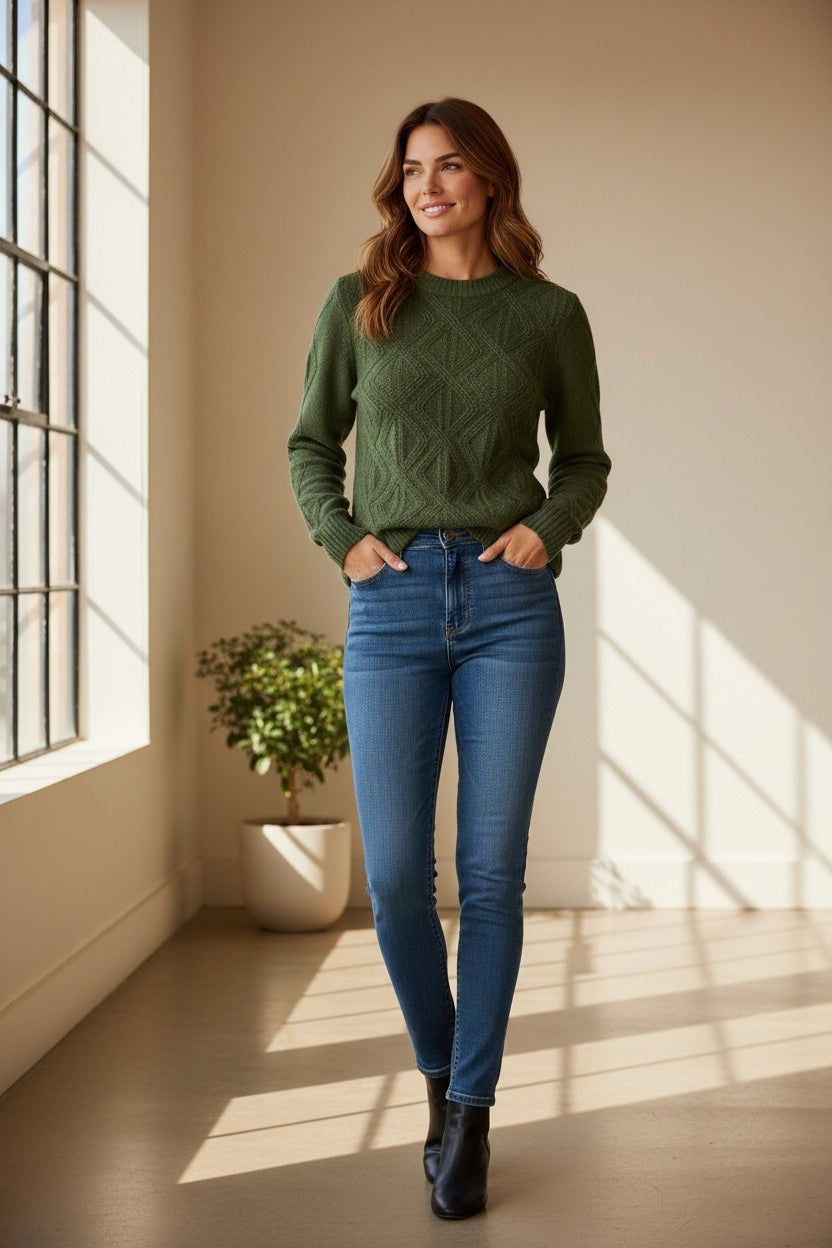 A woman stands indoors by large windows, smiling with her hands in her pockets. She wears the KINROSS CASHMERE - DIAMOND CREWNECK CASHMERE SWEATER in green from KINROSS, styled with blue jeans and black boots. Light pours through the windows, illuminating her luxurious layering piece. A potted plant sits in the background.