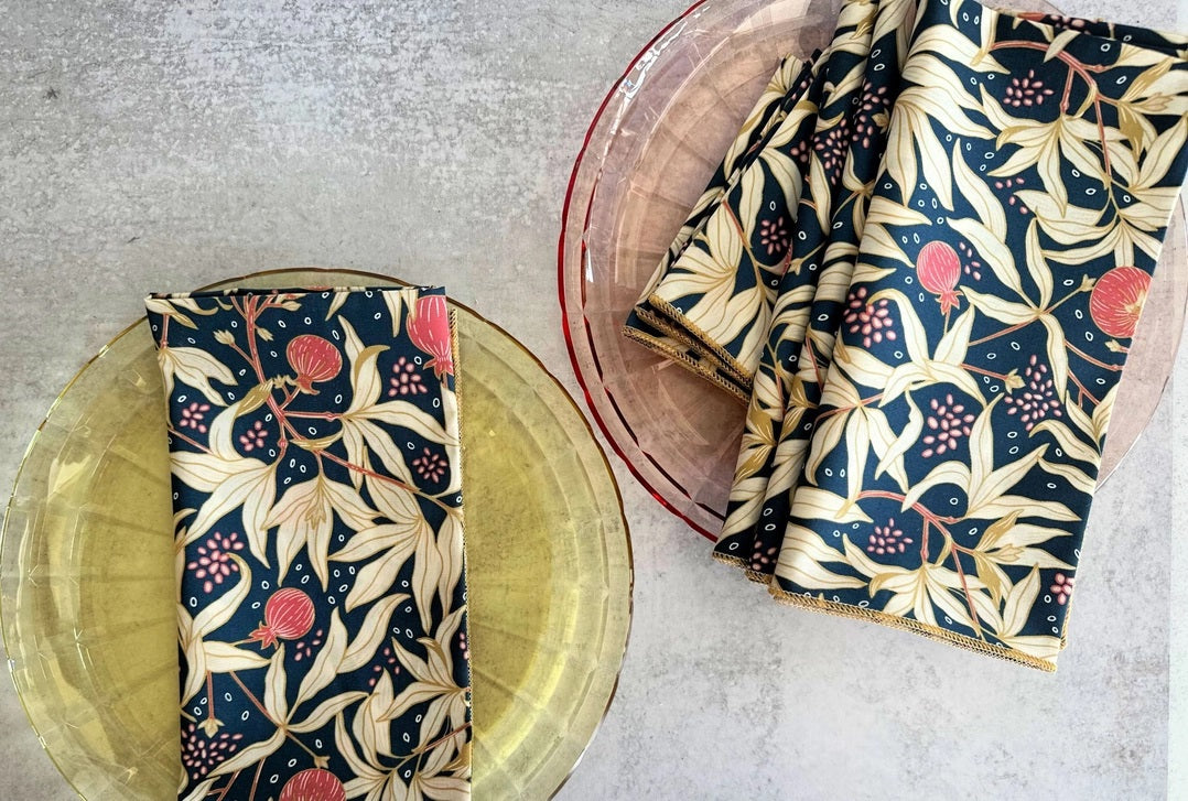 Two transparent plates, one yellow and one pink, rest on a light surface. Each displays DOT AND ARMY's Gooseberry Dinner Napkin—a dark blue cotton napkin with a beige, red, and green leaf pattern for stylish sustainable dining.
