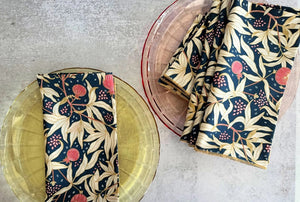 Two transparent plates, one yellow and one pink, rest on a light surface. Each displays DOT AND ARMY's Gooseberry Dinner Napkin—a dark blue cotton napkin with a beige, red, and green leaf pattern for stylish sustainable dining.