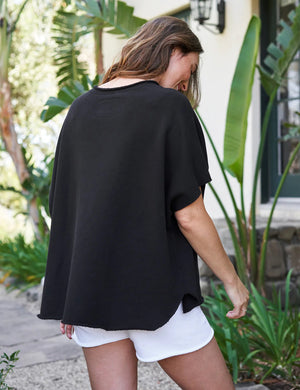 The FRANK & EILEEN OLIVE ORIGINAL CAPELET in black, by FRANK & EILEEN, is a black oversized T-shirt with a round neckline and short, wide sleeves. Made from 100% cotton, it features a loose fit and simple design reminiscent of a capelet against a light textured background.