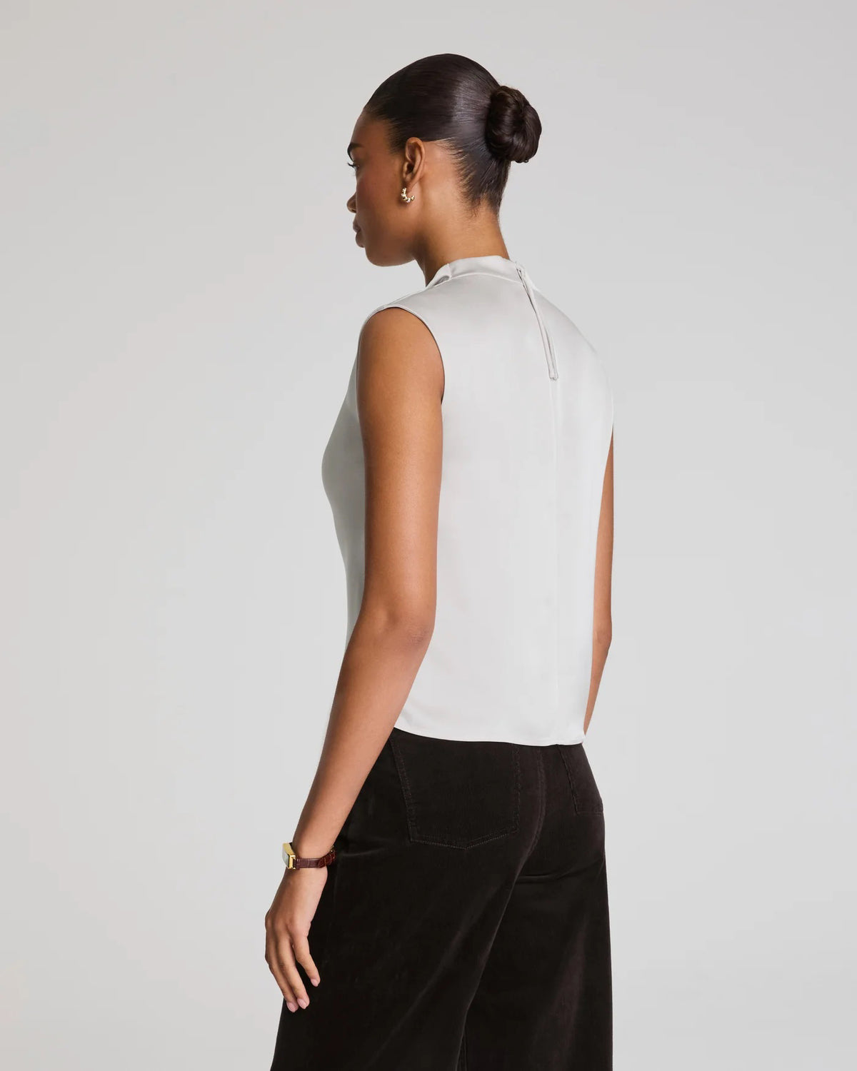 A woman wears the SPANX Silky Smooth Cowlneck Sleeveless Top in light gray with dark high-waisted pants, sitting on a stool against a plain background and looking at the camera with a neutral expression.