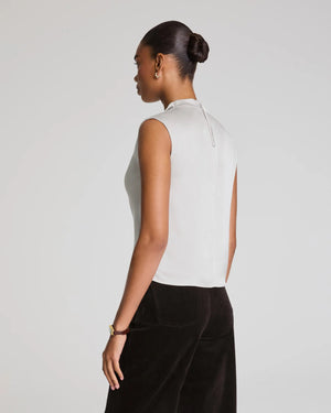 A woman wears the SPANX Silky Smooth Cowlneck Sleeveless Top in light gray with dark high-waisted pants, sitting on a stool against a plain background and looking at the camera with a neutral expression.