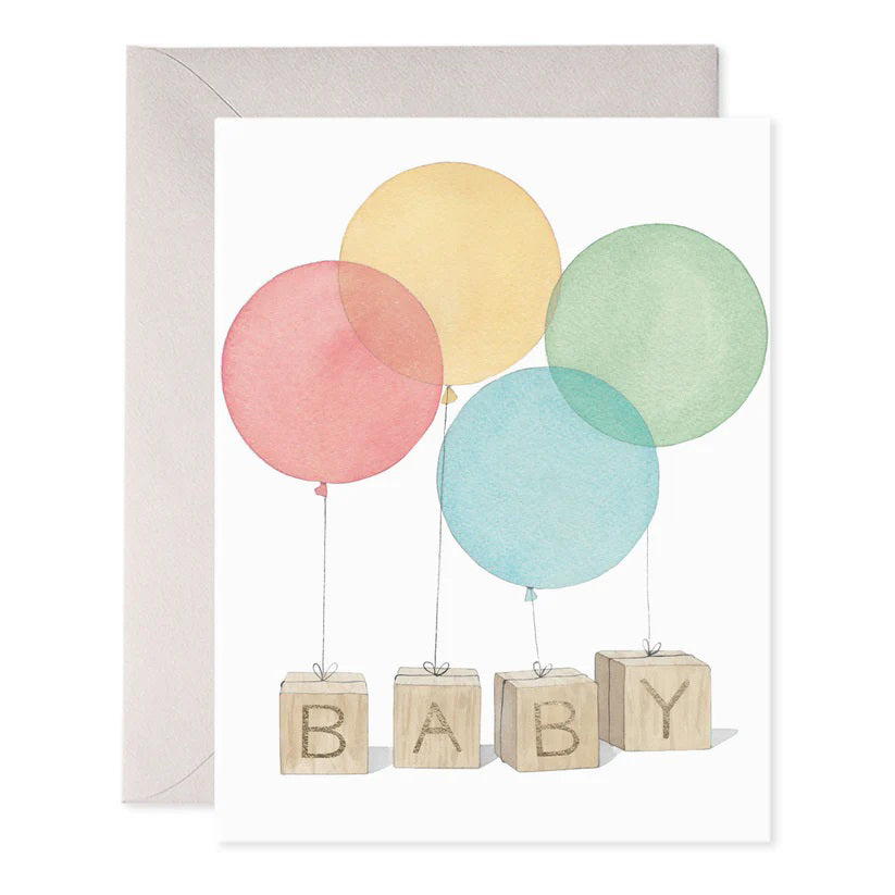 The E. FRANCES PAPER BABY BLOCKS GREETING CARD features four colorful balloons in red, yellow, green, and blue tied to wooden blocks spelling "BABY." This handcrafted card, made in the USA, comes with a plain envelope.