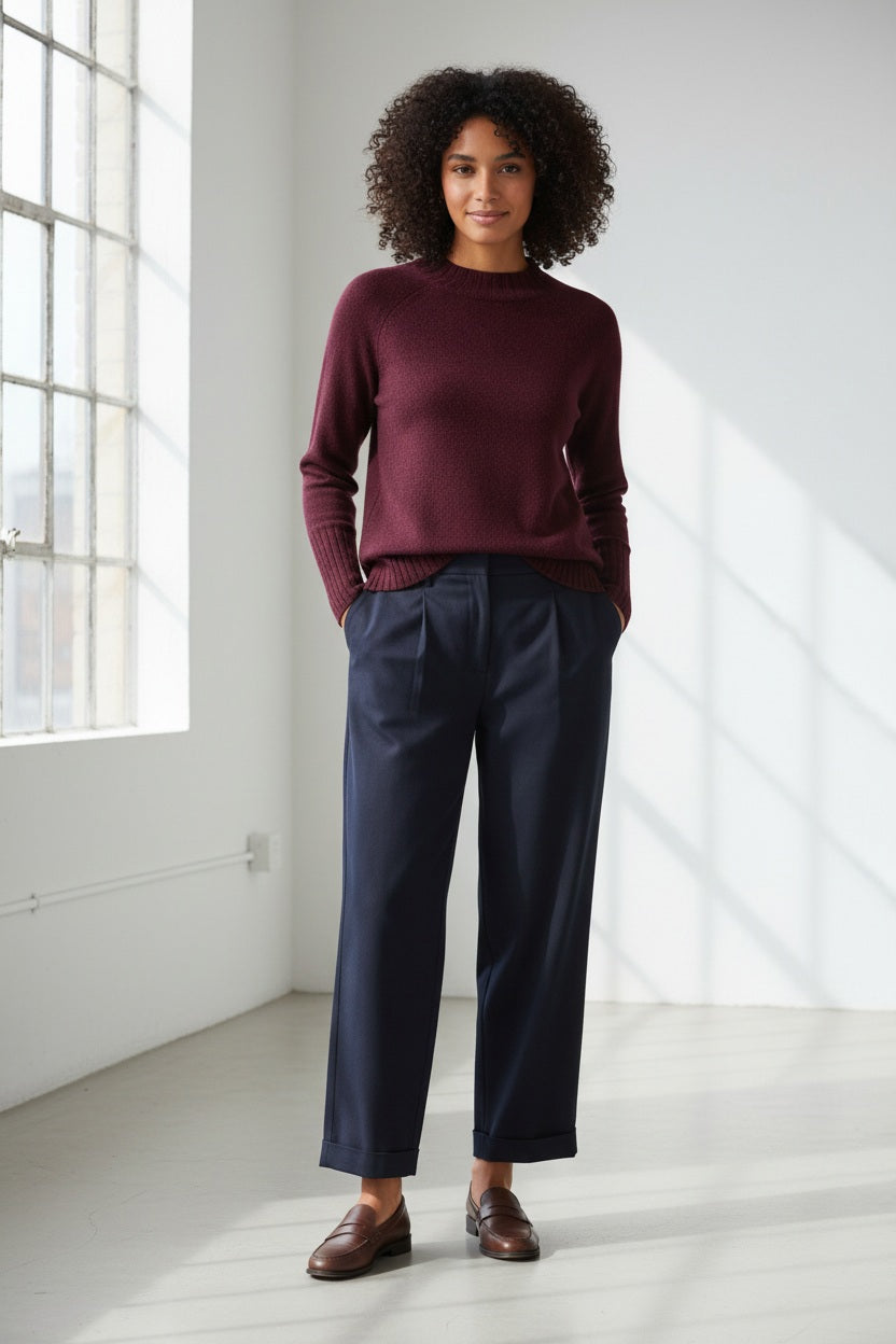 A person with long, light brown hair wears the KINROSS CASHMERE Hi Low Rib Trim Funnel Cashmere Sweater in Cordovan and dark pants, standing against a plain light background with one hand in their pocket, looking at the camera and smiling slightly.