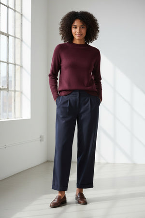 A person with long, light brown hair wears the KINROSS CASHMERE Hi Low Rib Trim Funnel Cashmere Sweater in Cordovan and dark pants, standing against a plain light background with one hand in their pocket, looking at the camera and smiling slightly.