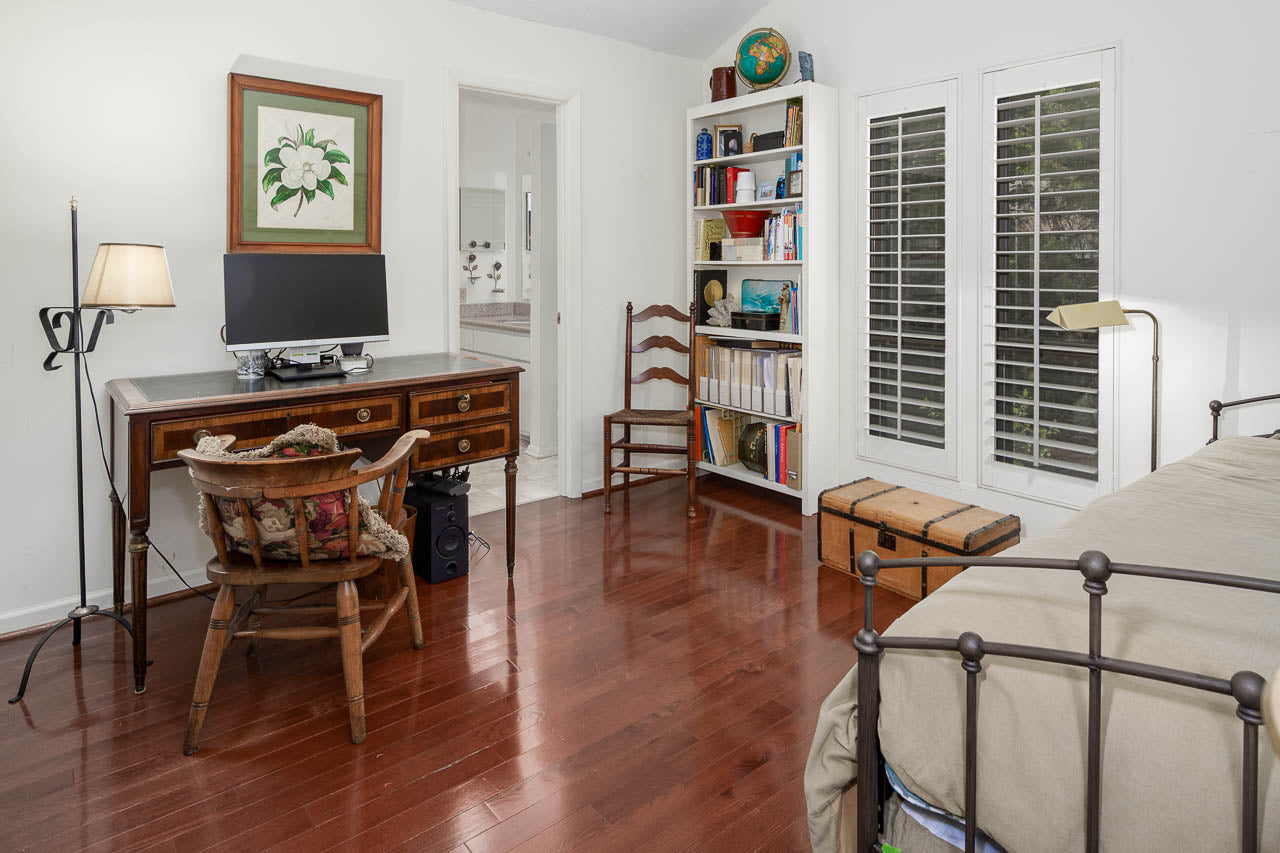 A cozy room with a wooden desk and computer, a wooden chair, bookshelves, a bed with a metal frame, hardwood floors, a lamp, and large windows with white shutters. There is artwork and a decorative globe on display.