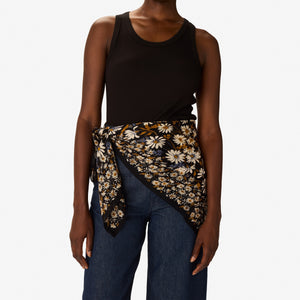 A woman with short hair, in a black sleeveless top and blue pants, stands against a white background. She wears the INOUI EDITIONS FAUST SCARF IN BLACK from INOUI, featuring yellow and white flowers, wrapped around her neck.
