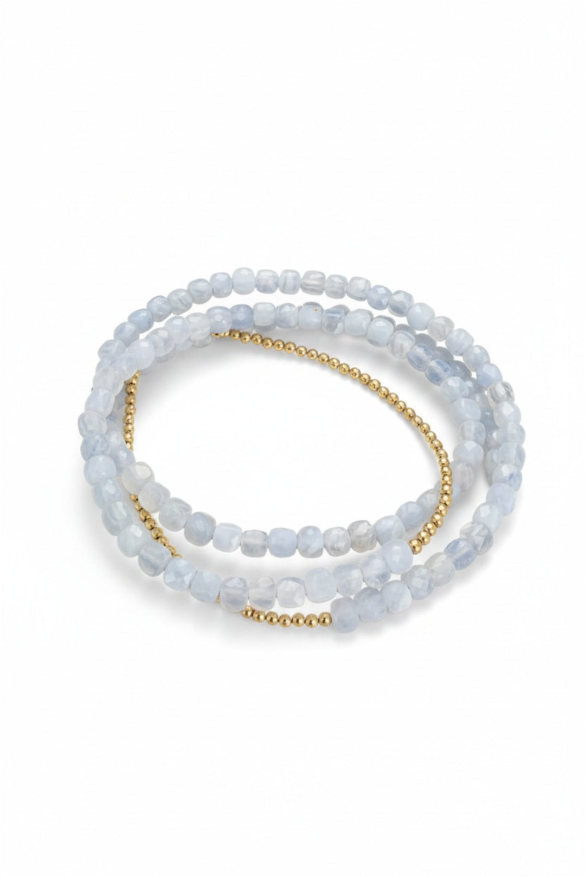 The ANN LIGHTFOOT Chalcedony Squares Triple Wrap Bracelet features two strands of light blue, translucent beads evoking the look of chalcedony, paired with a strand of small, shiny gold beads, all artfully coiled together against a white background.