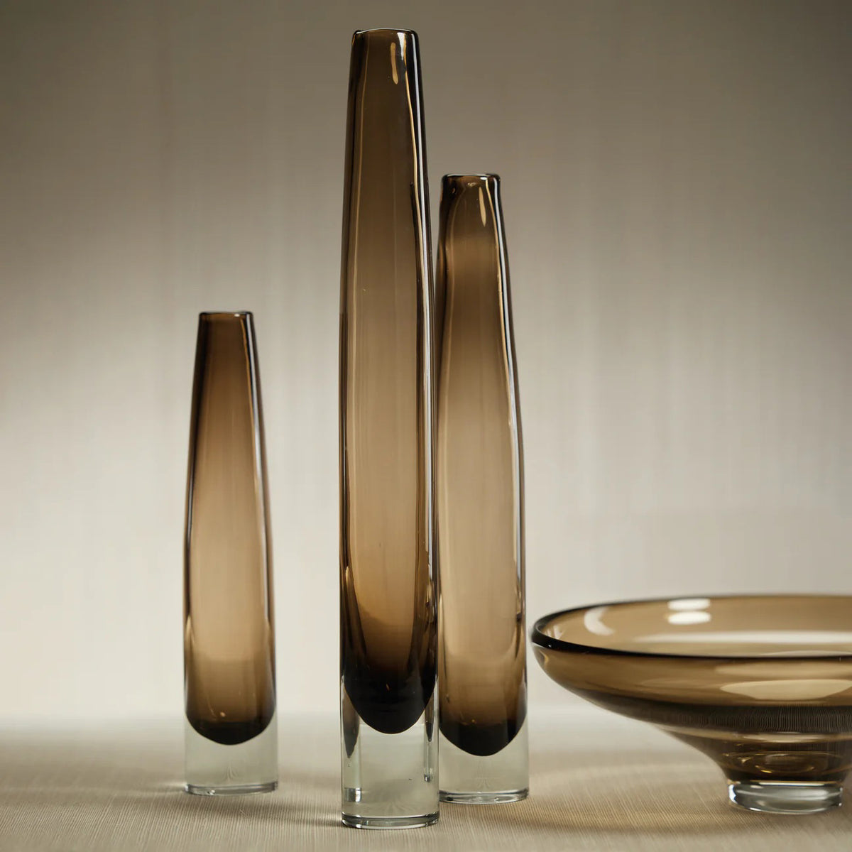 The ZODAX TATE SLIM GLASS TAUPE VASE - SHORT features a cylindrical taupe glass body with a clear base, measures 2.5" x 13.5", and is displayed against a plain white background.