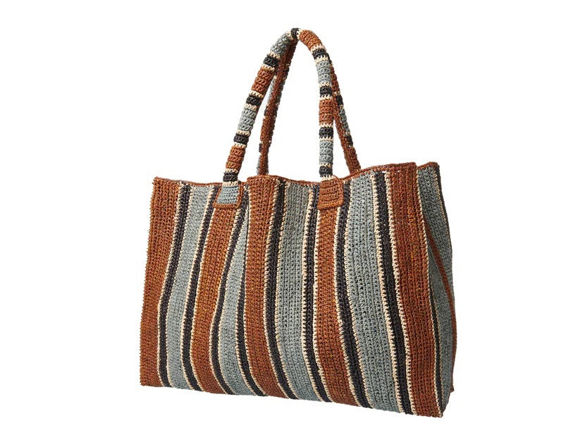 A woman stands on a sunlit path in a sleeveless dress and long scarf, holding the GABRIELE FRANTZEN - RAFFIA WEEKENDER BAG TOBACCO. She looks over her shoulder, surrounded by lush greenery.