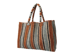 A woman stands on a sunlit path in a sleeveless dress and long scarf, holding the GABRIELE FRANTZEN - RAFFIA WEEKENDER BAG TOBACCO. She looks over her shoulder, surrounded by lush greenery.