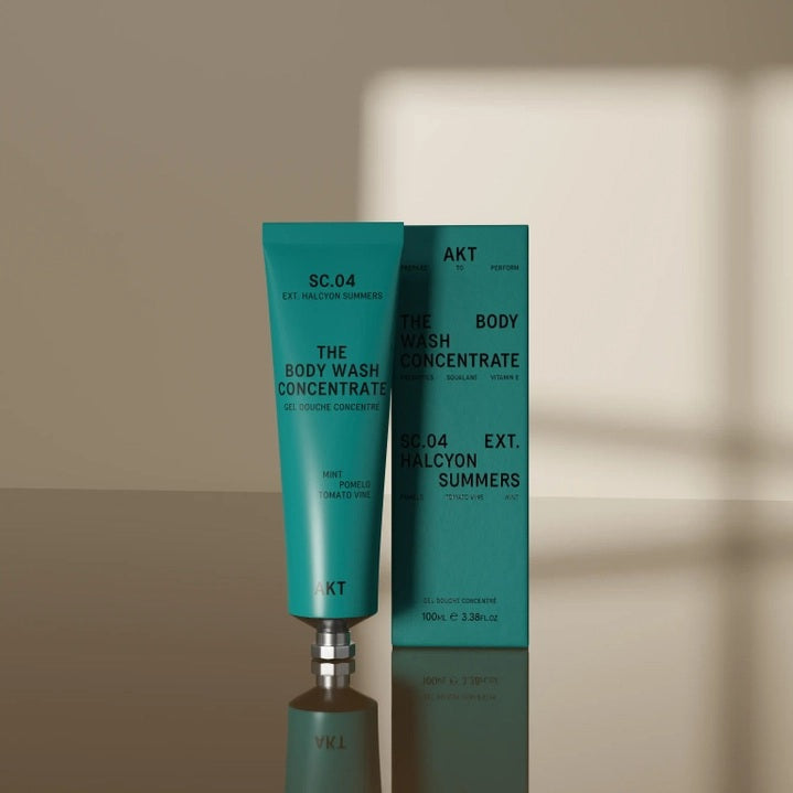 A person lathers up their back with a sponge against a teal backdrop, savoring the rich foam of AKT LONDON’s BODY WASH CONCENTRATE HALCYON SUMMERS. Three scent labels—Mint, Pomelo, and Tomato Vine—are displayed vertically on the right side of the image.