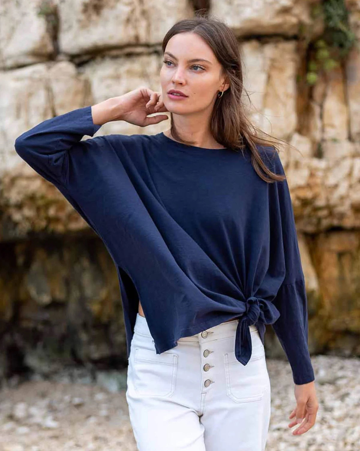 A woman with wavy brown hair models the MERSEA - CATALINA SLUB TEE—an oversized, long-sleeved white top with thin gray stripes—by MERSEA. She pairs it with blue jeans and stands against a plain, light background, hands behind her back.