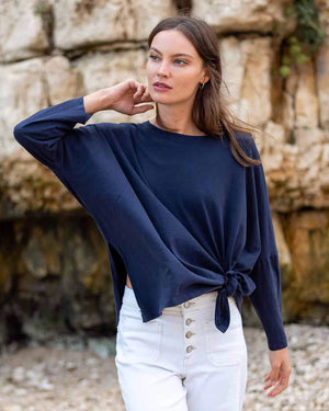 A woman with wavy brown hair models the MERSEA - CATALINA SLUB TEE—an oversized, long-sleeved white top with thin gray stripes—by MERSEA. She pairs it with blue jeans and stands against a plain, light background, hands behind her back.