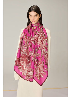 A person stands against a plain beige background wearing a long cream-colored sweater dress and the INOUI EDITIONS - ARBORIS WOOL SCARF IN PINK by INOUI, a bright pink, 100% wool scarf featuring large floral patterns and made in India.