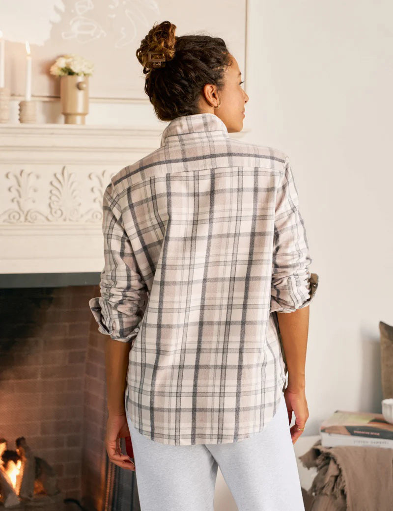 A woman smiles with her eyes closed, wearing the FRANK &amp; EILEEN EILEEN Relaxed Button Up Shirt in Italian Flannel Cream Sand Grey Plaid, paired with light gray sweatpants in a cozy room by a fireplace, with candles and books in the background.