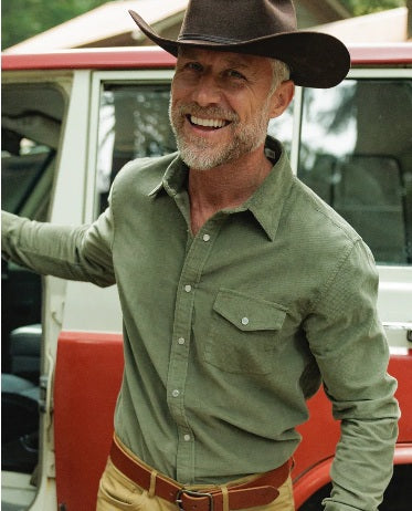 A smiling man with a gray beard, wearing the CRIQUET Long Sleeve Corduroy Pearl Snap Shirt in Olive and a brown cowboy hat, stands by the open door of a red and white vehicle.