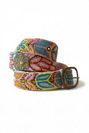 A person wears a cream-colored knit dress, a burnt orange cardigan, and a JENNY KRAUSS - FOLKLORICA FLORAL EMBROIDERED BELT IN CREAM with colorful floral embroidery and an oval buckle.