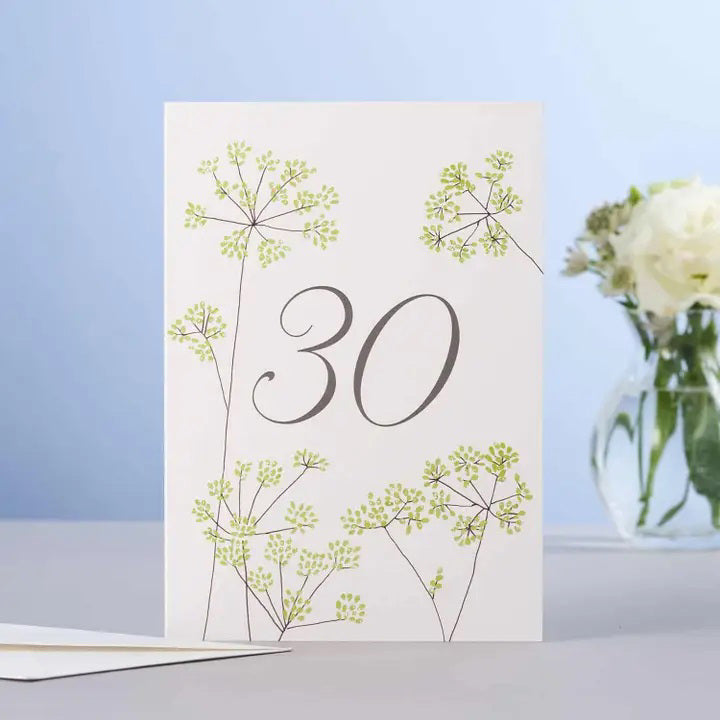 The ELOISE HALL LTD - FENNEL 30TH BIRTHDAY CARD features the number "30" on the front, encircled by intricate green floral designs. Crafted from premium GF Smith Board, this card is displayed on a light blue surface with its matching envelope beside it and a vase of white flowers in the background.