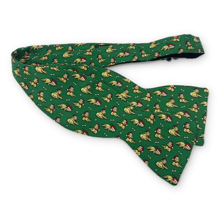 Add a touch of holiday cheer to your ensemble with the BIRD DOG BAY - SANTA PAWS SILK BOW TIE IN MID-GREEN. This playful, handmade bow tie by BIRD DOG BAY features a mid-green silk background adorned with a repeating pattern of yellow dogs wearing red hats, chewing on bones, and playing with balls—making it the perfect festive accessory for any holiday gathering.