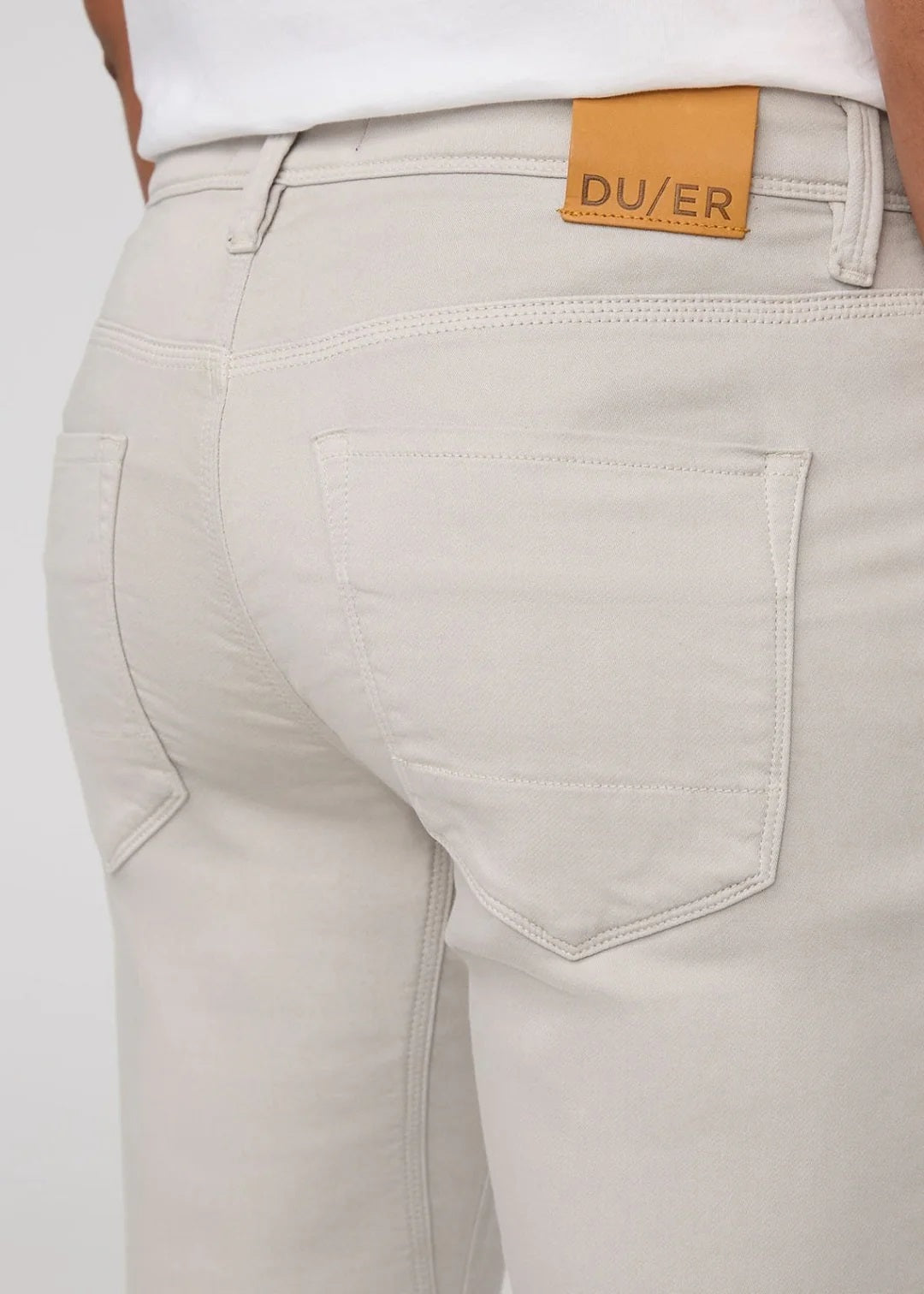 A person stands facing forward on a plain light background, wearing DUER NO SWEAT ATHLETIC TAPER PANTS IN MIST and matching light gray sneakers, feet slightly apart.
