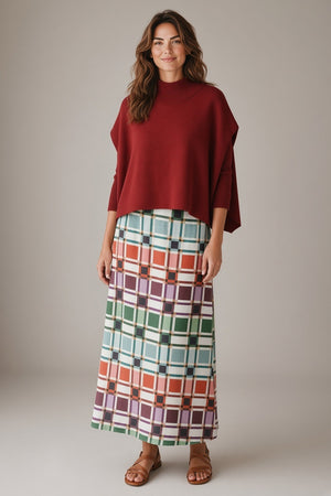 A woman wears a dark red knit poncho over a multi-colored BRITON COURT TIFFANY SKIRT IN CANDY BOX CHECK PLAID and brown sandals, perfect for a fall wardrobe.