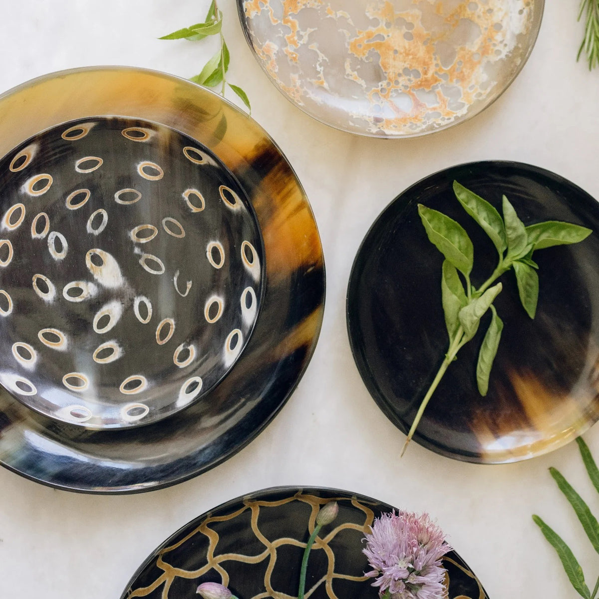 The BE HOME - SPLATTER HORN PLATE SMALL by BE HOME, INC is a round, shallow plate handmade in Vietnam from water buffalo horn, showcasing a dark brown and amber marbled pattern on a beige surface.