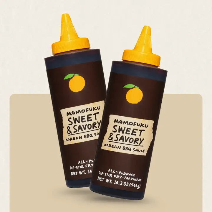 Two bottles of MOMOFUKU - SWEET & SAVORY KOREAN BBQ SAUCE by MOMOFUKU, each 16.3 oz, with yellow caps, dark labels, and an orange fruit logo. This all-purpose sauce offers a savory-sweet flavor that’s perfect for use as a dip, stir-fry ingredient, or galbi marinade.