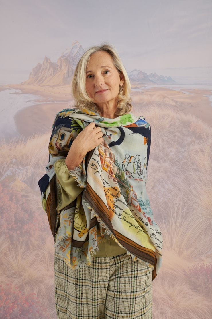 A woman with wavy blonde hair smiles while looking down. She wears a loose, light lavender blouse, dark pants, and the STORIATIPIC EUGENIE WOOL SCARF. Behind her is a scenic landscape of mountains, grass, and water.