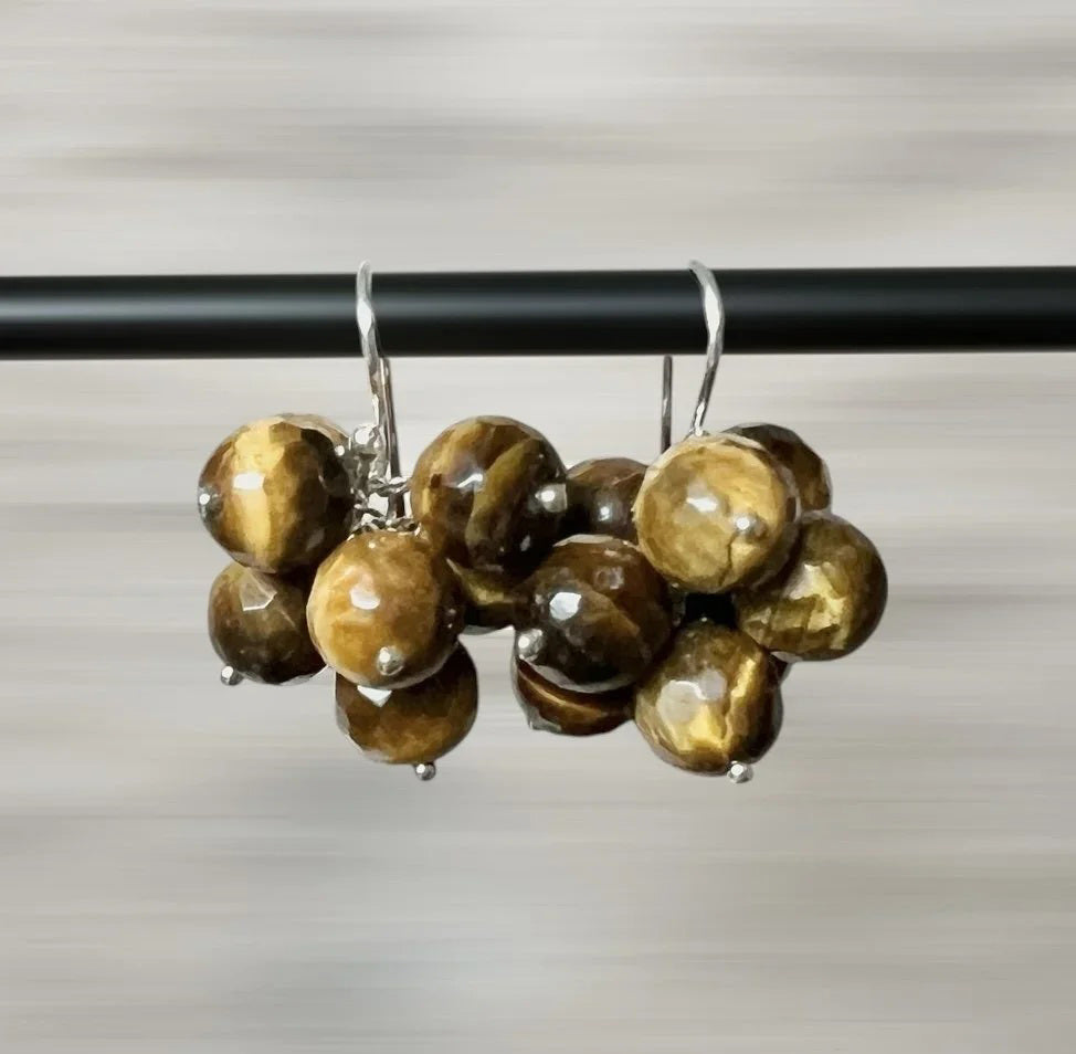 ANN LIGHTFOOT - TIGER EYE CLUSTER EARRINGS feature faceted round brown and golden tiger eye beads on sterling silver hooks, shown on a black rod with a blurred background.