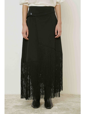 A person stands against a plain background wearing the MEIMEIJ - FRINGE WRAP SKIRT with an asymmetric hem, paired with a light top and black boots.