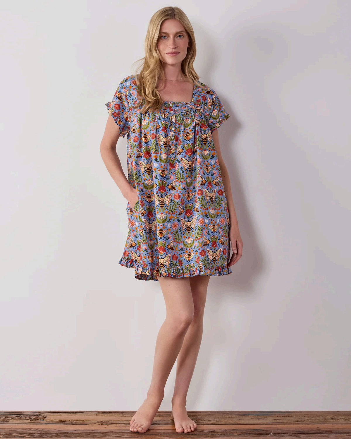 A woman with long blonde hair wears the PRINTFRESH Bee Tapestry Pinktuck Nightgown in Delphinium Blue, standing against a plain light-colored wall and smiling slightly.