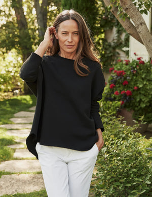 A woman stands outdoors in a garden, wearing a loose black long-sleeve top and white pants, with one hand touching her hair. Sunlight filters through trees and flowering plants are visible in the background.