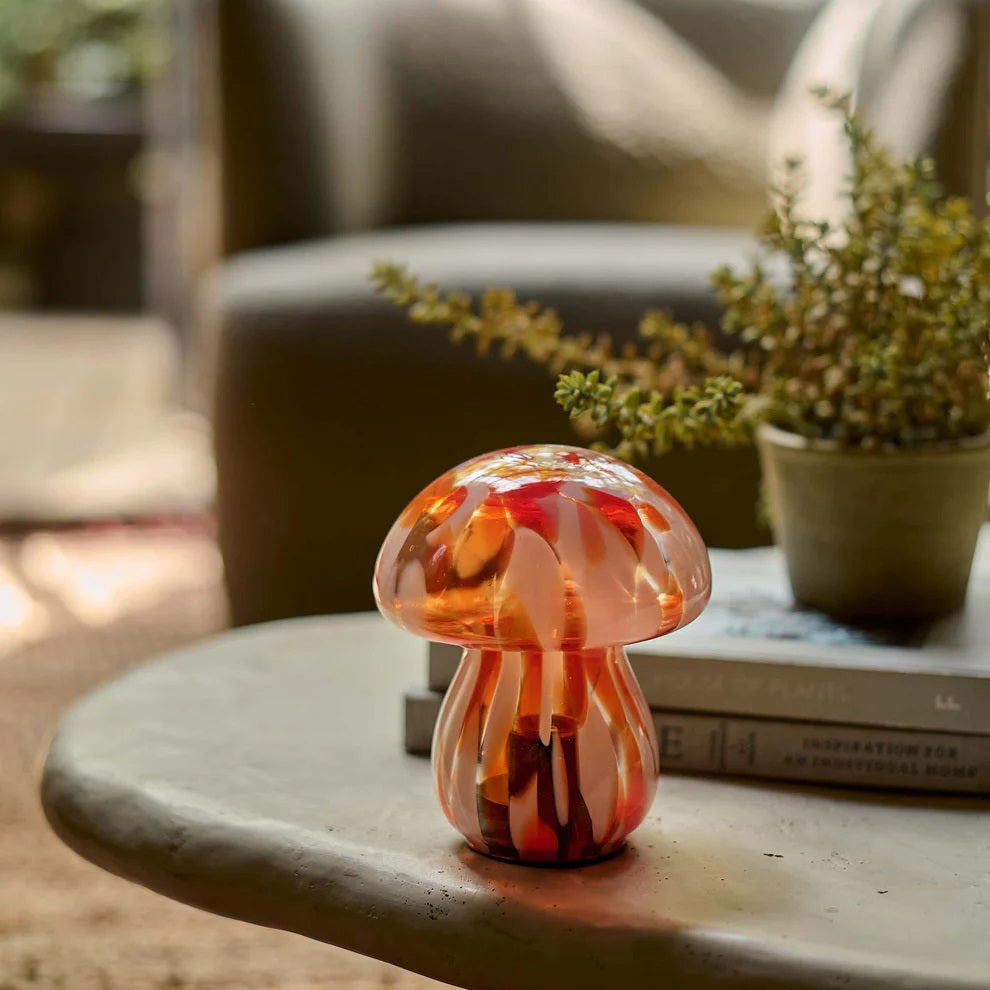 The ABIGAIL AHERN - MUSHROOM CORDLESS LED is a small glass lamp with a rounded cap and stem, featuring swirled pink, orange, and red hues—resembling a charming mushroom figurine against a plain white background.