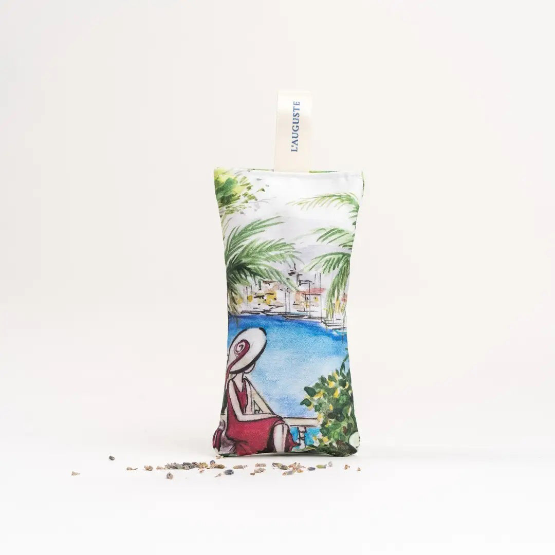 The L&#39;AUGUSTE PROVENCE - FRENCH LAVENDER SACHET FROM PROVENCE is a small, handcrafted sachet featuring a hat-wearing woman by the water with palm trees, filled with organic lavender and labeled “L&#39;AUGUSTE PROVENCE” for lasting freshness.