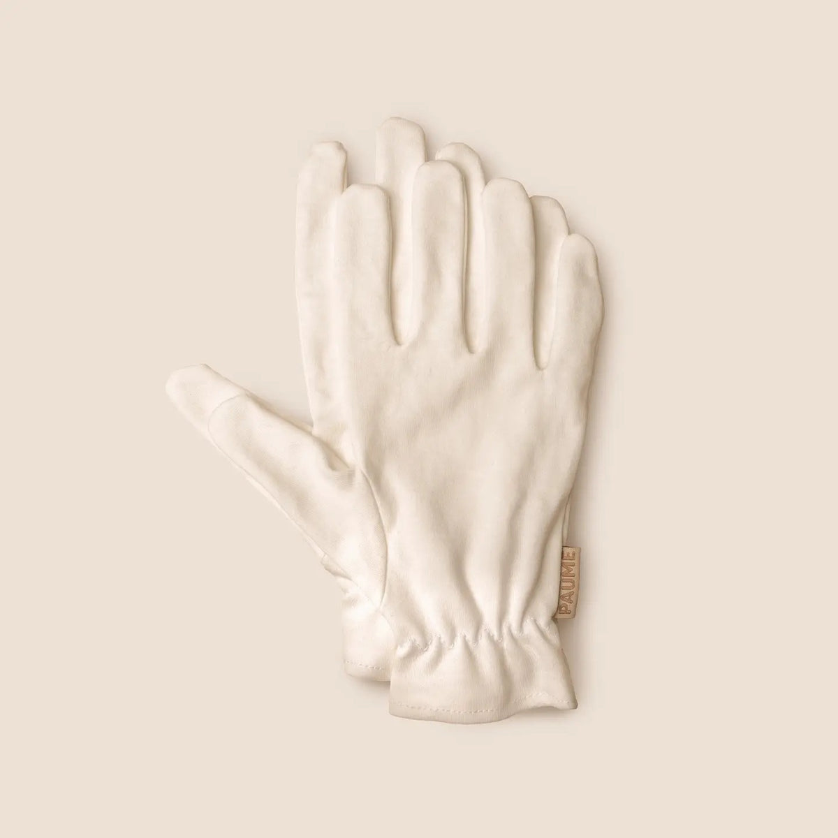 PAUME - ORGANIC COTTON OVERNIGHT GLOVES by PAUME are soft, off-white gloves with a subtle sheen, elastic wrists, and a simple design. Made from organic cotton, they’re perfect for nighttime moisturizing and shown on a matching beige background.
