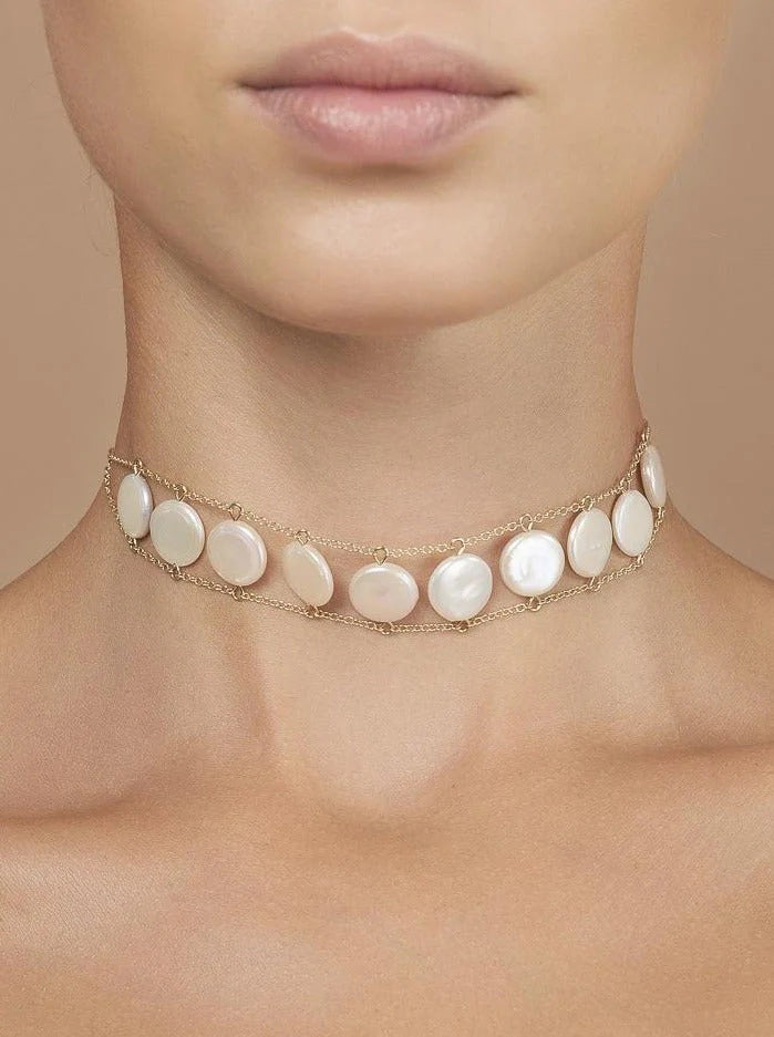 A close-up of a woman&#39;s neck wearing the GOLDISH Ladders to Bliss Pink Moonstone Choker, featuring two rows of round moonstones set in gold-toned chain links, by GOLDISH, against a neutral background.