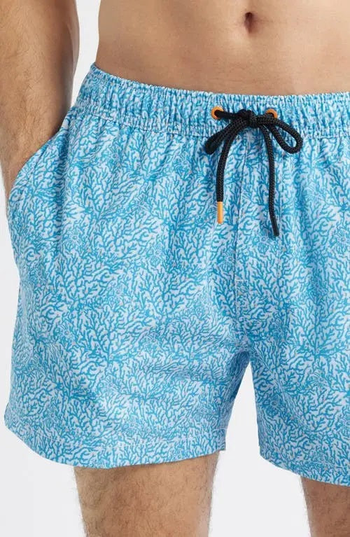A shirtless man wears SAVE THE DUCK - ADEMIR swim trunks in light blue corals NO FISHING, made from quick-dry recycled polyester with a black drawstring, standing against a plain white background.