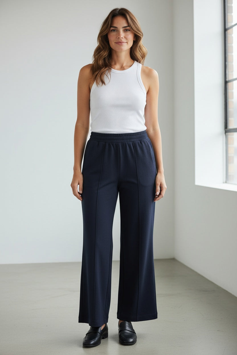 A person is standing sideways against a plain white wall, wearing a black long-sleeved mesh top, the MEIMEIJ STRIGHT LEG PULL ON PONTE PANT with a slight flare, and black flat shoes. The fingers appear to be partially covered by the sleeves of the mesh top.