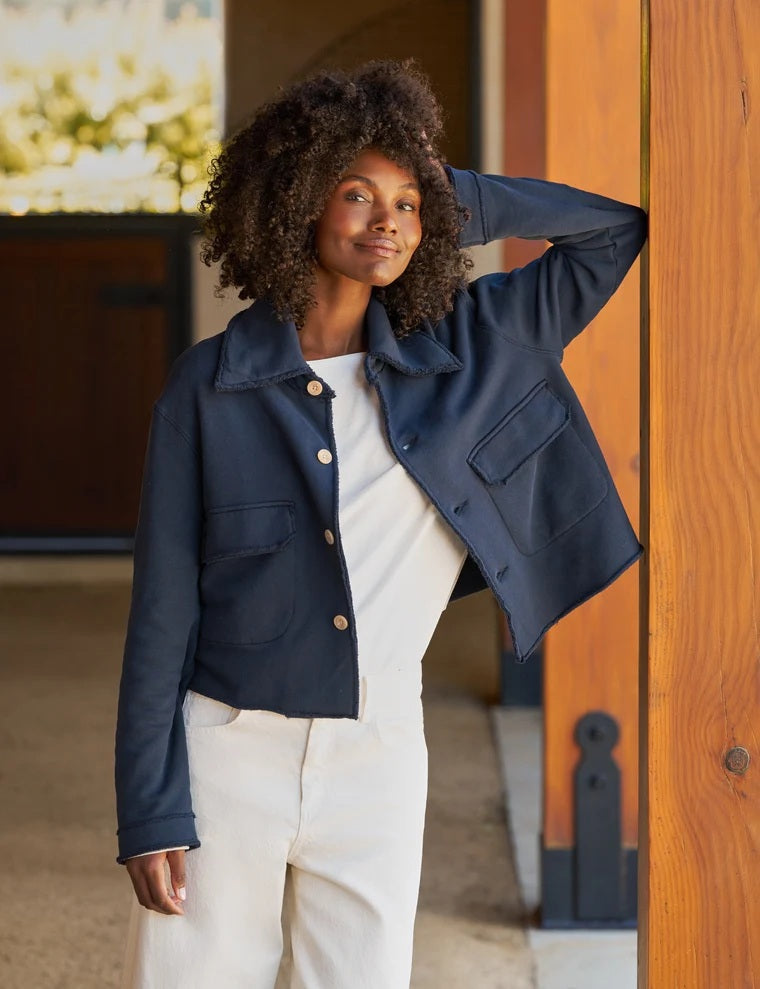 A woman with long brown hair wearing the FRANK & EILEEN Rowan Cropped Sweatshirt Utility Jacket in Triple Terry Classic Navy, paired with white pants, stands sideways and smiles outside near a white building with windows.