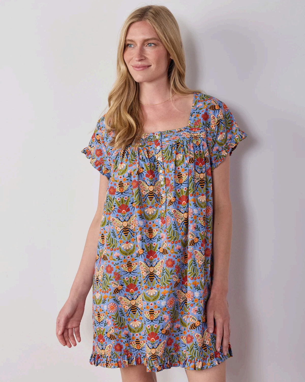 A woman with long blonde hair wears the PRINTFRESH Bee Tapestry Pinktuck Nightgown in Delphinium Blue, standing against a plain light-colored wall and smiling slightly.