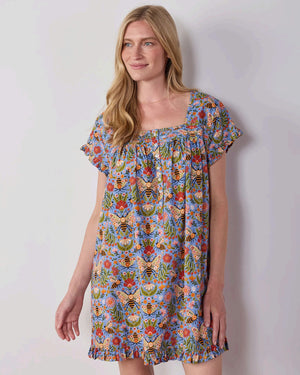 A woman with long blonde hair wears the PRINTFRESH Bee Tapestry Pinktuck Nightgown in Delphinium Blue, standing against a plain light-colored wall and smiling slightly.