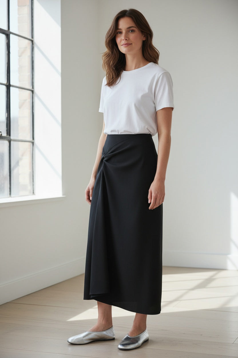 A woman stands indoors by a window, smiling softly in a white short-sleeve t-shirt and the MEIMEIJ SIDE GATHER SKIRT IN BLACK, featuring a knot detail in soft scuba fabric, her silver flats gleaming.