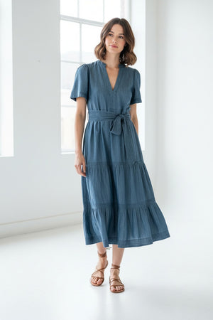 A woman with blonde hair, wearing the MERSEA Pariscope Maxi Dress in Chambray, stands by a stone wall. The blue denim maxi dress from MERSEA features short sleeves, a belted waist, and a subtle ruffle neckline for added elegance. She accessorizes with a gold necklace featuring a round pendant.