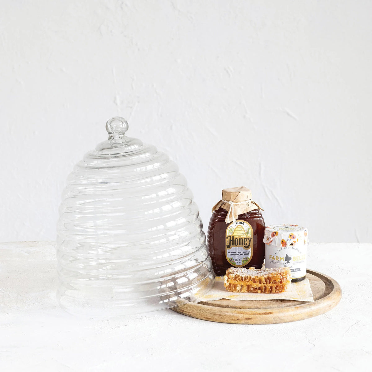 Introducing the BEE HIVE CLOCHE WITH TRAY by CREATIVE COOP: This eco-friendly serveware piece features a glass cloche shaped like a beehive with grooves resembling hive layers and now includes a sleek mango wood base. It comes with a spherical knob on top for easy lifting, and the cloche itself is made from reclaimed glass for added sustainability.