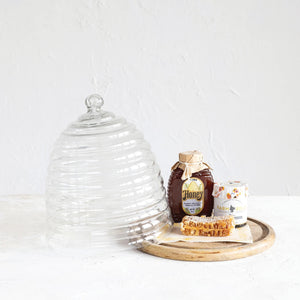 Introducing the BEE HIVE CLOCHE WITH TRAY by CREATIVE COOP: This eco-friendly serveware piece features a glass cloche shaped like a beehive with grooves resembling hive layers and now includes a sleek mango wood base. It comes with a spherical knob on top for easy lifting, and the cloche itself is made from reclaimed glass for added sustainability.