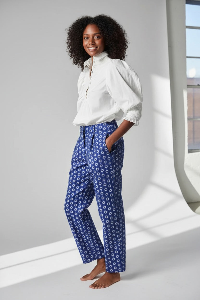 Rear view of a person wearing the Hope for Flowers Smocked Shirt, a bright red blouse with puffed sleeves, paired with blue jeans featuring pom-poms. Made from GOTS Certified organic cotton, this outfit embodies sustainable fashion against a gray background.