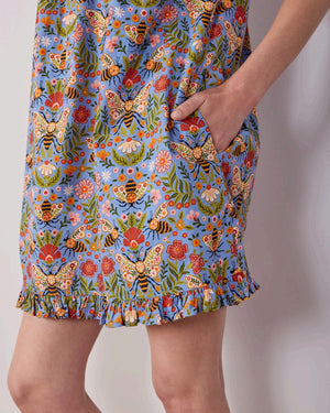 A woman with long blonde hair wears the PRINTFRESH Bee Tapestry Pinktuck Nightgown in Delphinium Blue, standing against a plain light-colored wall and smiling slightly.