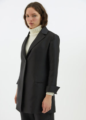 A person with long, wavy hair is wearing the BLACK BLAZER / JACKET by ALYKI LABORATORIO, made in Italy, over a white turtleneck sweater. They are standing against a white background with their arms crossed and have a serious expression on their face.