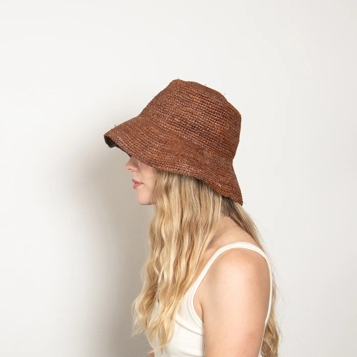 A woman with long, wavy blonde hair wears a white tank top and the WYETH HATS - TALI Raffia Bucket Hat in Chocolate by WYETH, shown in profile against a plain light background.